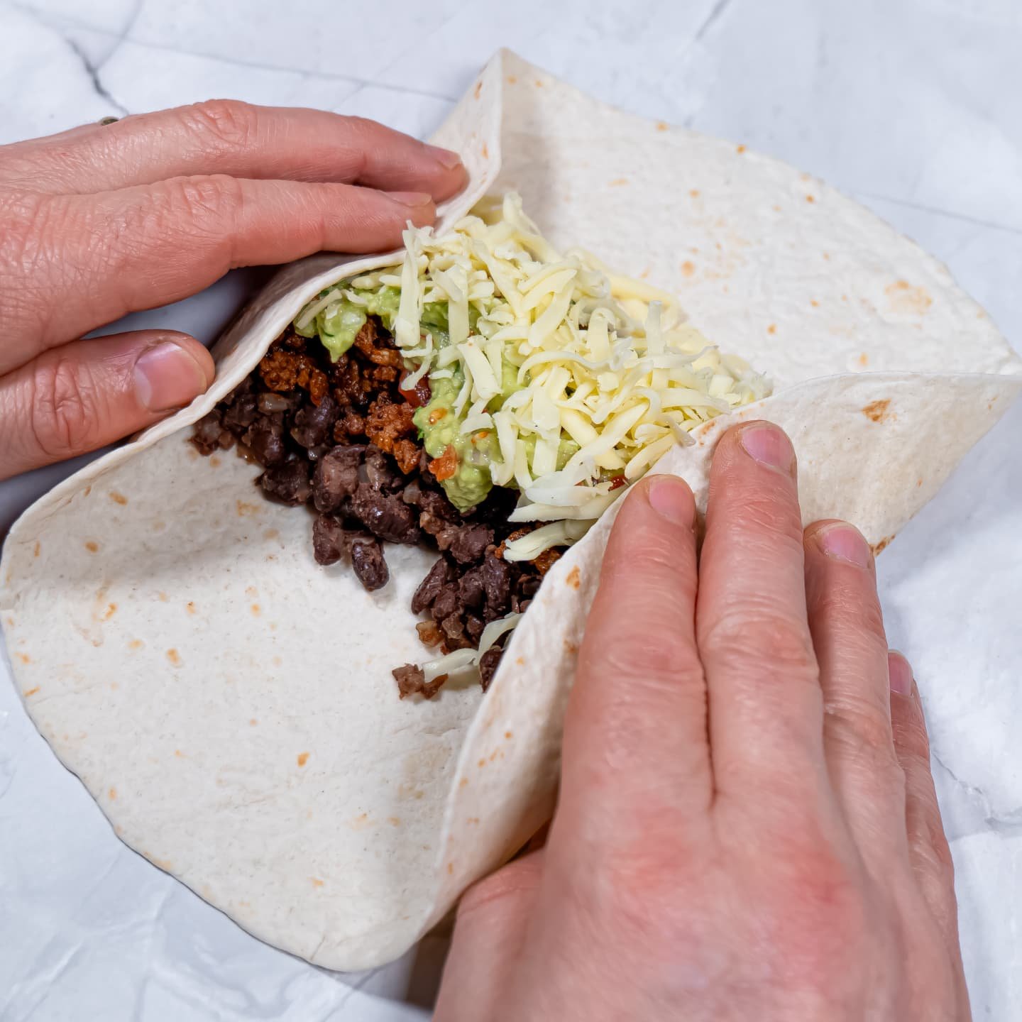 Hands folding the sides of a tortilla filled with seasoned beef, black beans, guacamole, and shredded cheese to prepare a burrito.