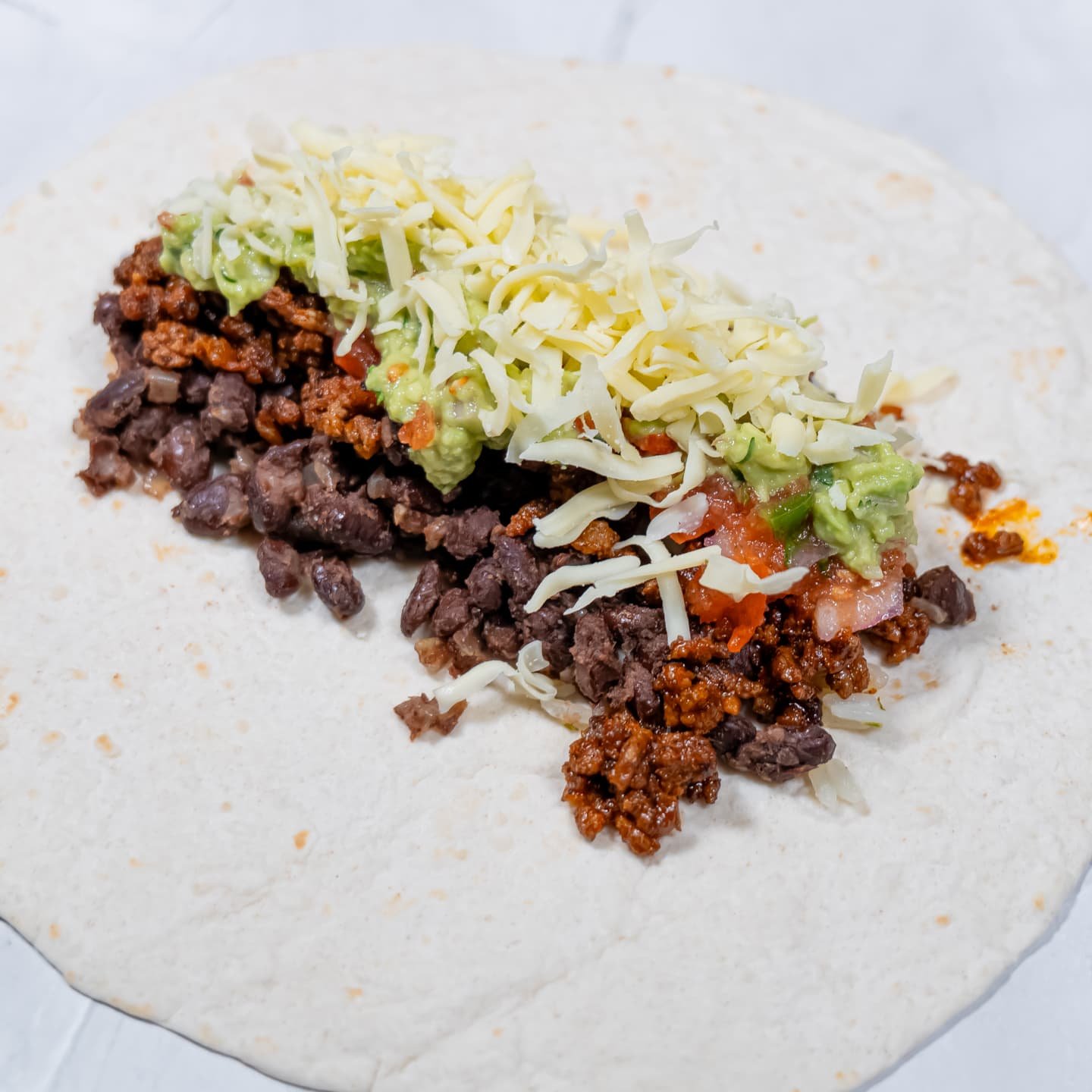 A flour tortilla with layers of seasoned beef, black beans, salsa, guacamole, and shredded cheese, ready to be wrapped into a burrito.