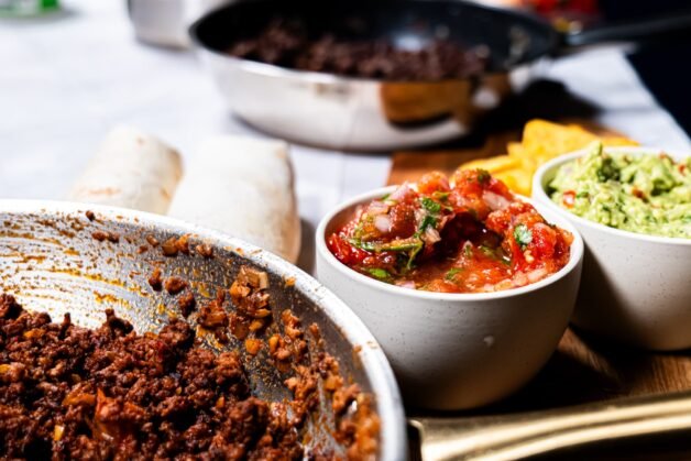 A skillet of seasoned ground beef with bowls of fresh salsa and guacamole, served alongside warm tortillas, ready for making burritos.