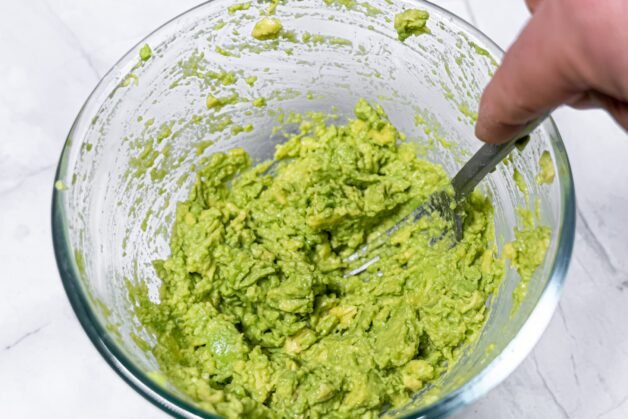 Mashing ripe avocados in a glass bowl with a fork for homemade guacamole.